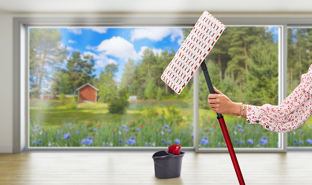 En arm i en sommerbluse holder en Vileda-moppe med en sommereng i baggrunden.